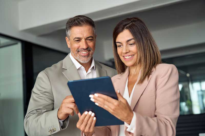 Smiling busy middle aged business man and business woman professional corporate executive leaders wearing suits discussing digital strategy using tablet