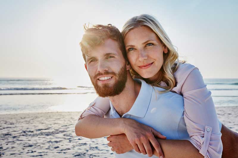 couple and man carrying woman in piggy back while they smile at the beach